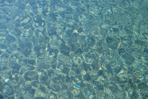 Background, closeup and texture of a blue water surface over a bottom of many pebbles. The sun shines on the fresh water.