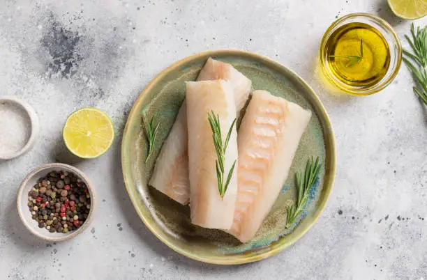 Ceramic plate with fresh cod fish, rosemary, lime, salt and olive oil on gray table. Mediterranean cuisine. copy space