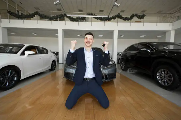 Photo of Young man in a suit is on his knees in a car dealership and is happy to buy a new car.
