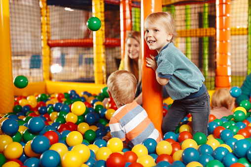 Kids cheering and playing with plastic colourful balls. Happy kids playing in amusement park balls pool with young teacher. Children throwing balls high. Playing with balls: activities for children