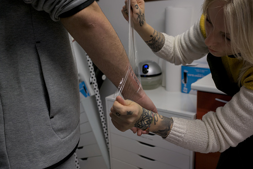 white-skinned tattoo artist applies the transparent protective film to the newly tattooed arm of the young client