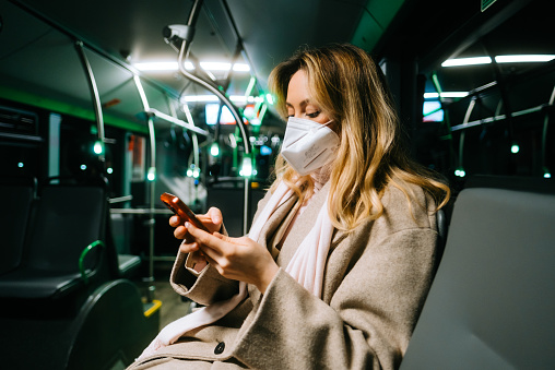 An attractive caucasian woman in protective mask using a smartphone while riding a bus in the night. Young beautiful woman using public transportation during covid.