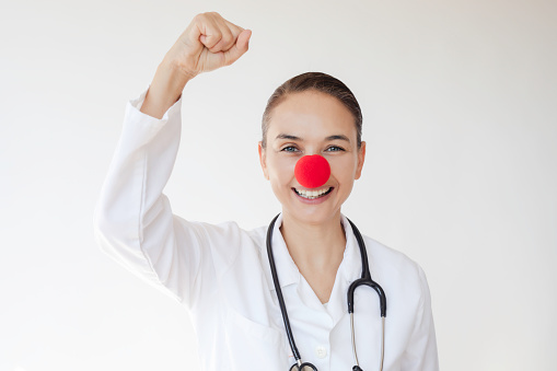 Doctor with red clown nose is punching the air with a toothy smile in front of white background.