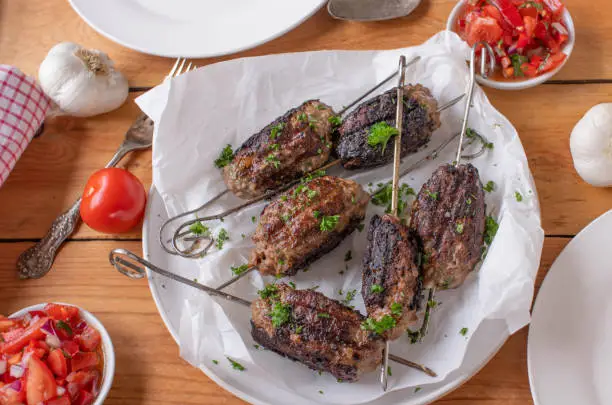 Spicy grilled minced meat skewers, cevapcici or mititei served with fresh tomato salad on wooden table. Top view. Delicious barbecue meal for summer