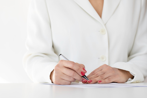 Unrecognizable caucasian woman wearing white jacket and red nail polish is signing contract paper.