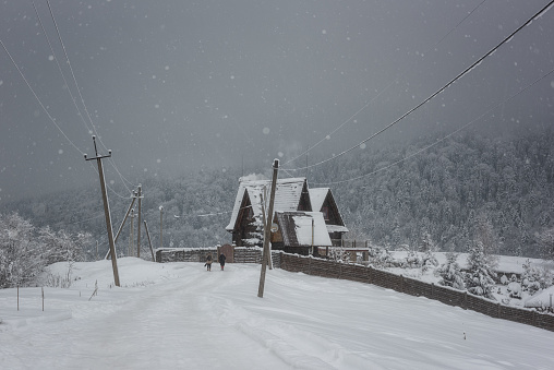 Snowfall in the mountain countryside, rural winter landscape with white fluffy snow, wooden house, snowflakes and snowy mountains on background, extreme travel