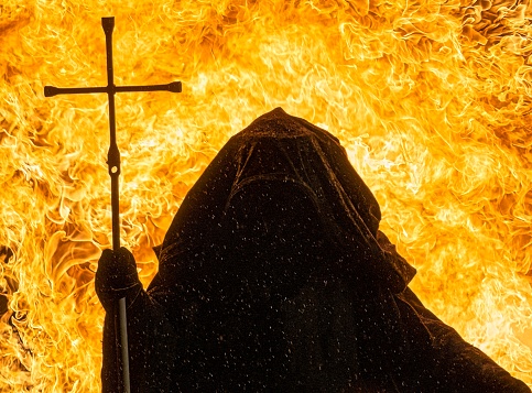 A man in hood holding a steel cross with fire background
