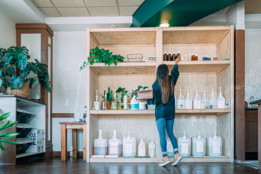 Cheerful college age woman standing in front of a display of sustainable self care products in a zero waste store. She is stocking shelves with reusable glass bottles available for sale. The store, located in the United States, is filled with a large group of self care items, including body wash, shampoo, and other products, all arranged in an attractive and visually appealing way. This photo represents a sustainable lifestyle, with a focus on environmental conservation and ethical consumerism. It is suitable for use in a variety of contexts, including retail, finance and the economy, and social issues.