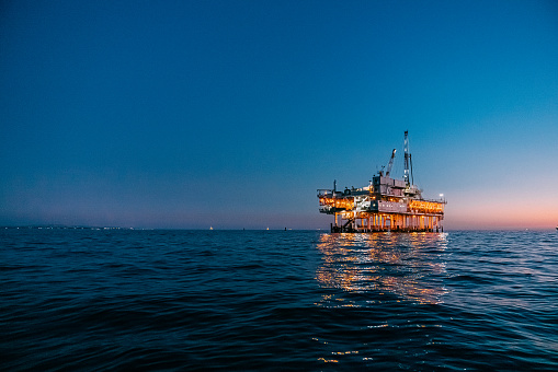 A beautiful image of an offshore oil rig at dusk off the coast of Huntington Beach, California. The orange tones of the setting sun highlight the industrial machinery and equipment used in the drilling and extraction of fossil fuels, including crude oil and natural gas. 

This photograph captures the intersection of the energy industry and the natural beauty of the Pacific Ocean, and speaks to issues of fuel and power generation, energy crises, and environmental concerns surrounding the oil and gas industry.