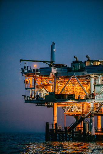 A stunning photograph of an offshore oil rig at dusk off the coast of Huntington Beach, California. The orange, pink, and purple tones of the setting sun highlight the industrial machinery and equipment used in the drilling and extraction of fossil fuels, including crude oil and natural gas. \n\nThis image captures the intersection of the energy industry and the natural beauty of the Pacific Ocean, and speaks to issues of fuel and power generation, energy crises, and environmental concerns surrounding the oil and gas industry.\