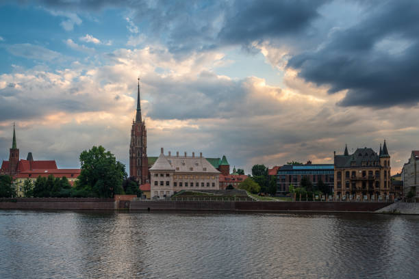 The photograph captures the stunning view of the Cathedral on Ostrow Tumski, the oldest part of Wrocław, Poland, from the banks of the Oder River at dawn. The early morning light illuminating the facade of the Cathedral, and the calm waters of the river in the foreground, creates a peaceful and serene atmosphere. The photo showcases the beauty of the historical architecture and the natural landscape that can be found in Ostrow Tumski, and how they are harmoniously blend together. The Ostrow Tumski's Cathedral is known for its architectural and cultural significance as well as tourist attraction.