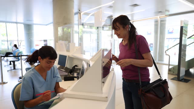 Female patient arriving to the hospital for a medical appointment and receptionist handing her a form to fill in