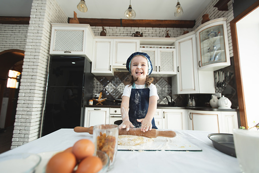 A little girl in an apron and a chef's hat is rolling out cookie dough, laughing