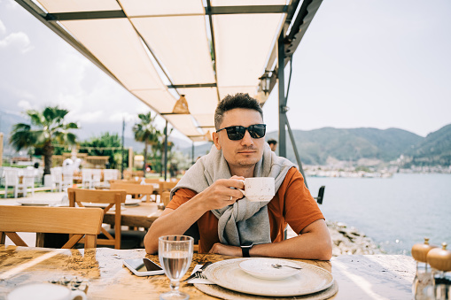 Young man male in sunglasses drinking coffee in a street outdoors seaside cafe restaurant coffee shop with scenery sea and mountains in the background. Hello summer holiday vacation.