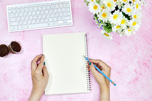flat lay of workplace : woman holding a blue pencil and writing on empty notebook, keyboard and muffins and daisy flowers