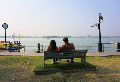 Kolkata, WB. India - December 11, 2022
Rear View of a couple sitting on a bench facing the lake at Eco Park. Sunny day.