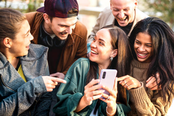 Multicultural young people having fun with mobile phone at urban city yard - Happy guys and girls spending time together sharing funny content online on smartphone social networks - Bright warm filter stock photo