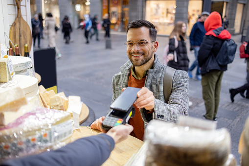Happy young adult man buying cheese from a local cheese vendor in the city, paying for it using his smart phone