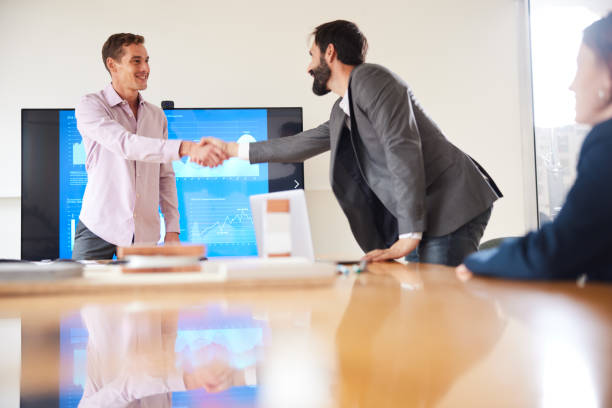 Male business presenter greets a new arrival with a handshake over the conference table