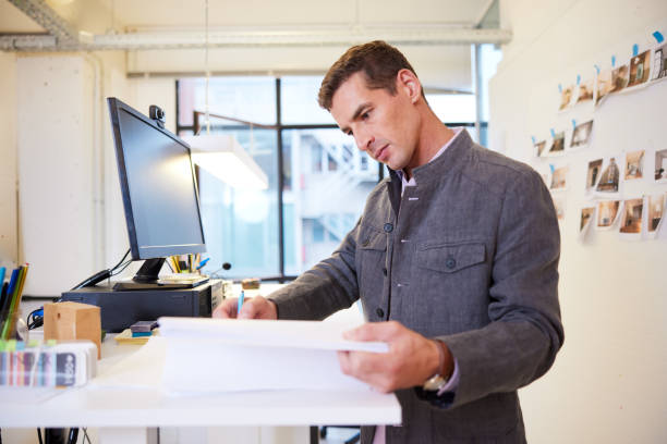 istock Mid adult business  standing and working at a flexible work desk with desktop PC 1455543758