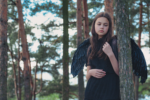 Teenage girl with long hair in black wings posing in fairy tale woodland, closed eyes. Young pretty lady Princess angel at mysterious forest. Vintage retro image, fantasy concept. Copy text space