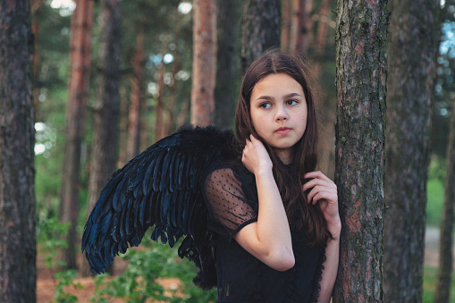 Portrait teen girl with long hair in black wings posing in fairy tale woodland, looking away. Young lady Princess angel at mysterious forest. Vintage retro image, fantasy concept. Copy text space