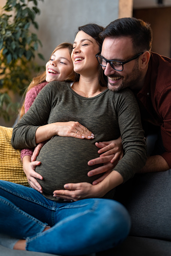 Smiling pregnant mother having an emotional support during pregnancy from her husband and daughter. Happy healthy family embracing at home, excited about the new family member.