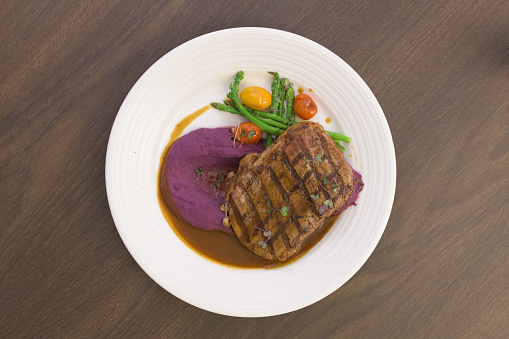 Beef steak on a white plate with grilled tomatoes, asparagus, mashed purple sweet potato and brown sauce.