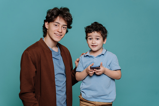 Handsome caucasian guy explains to younger brother how to use phone, two brothers standing against turquoise studio background. Cute curly baby boy holds smartphone enjoys game. Brotherhood.