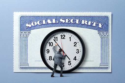 A man holds back the hands of time on a large clock that stands in front of a large Social Security card.  The image conveys the issues of the solvency of the social security system or a person's invetibile march toward their eligiblity for social security.