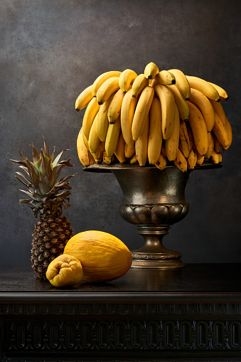 A  pinepple, yellow fruit, large silver vase and a bunch of bananas on an antique table with a black stone wall in the background.  Very detailed and sharp photography