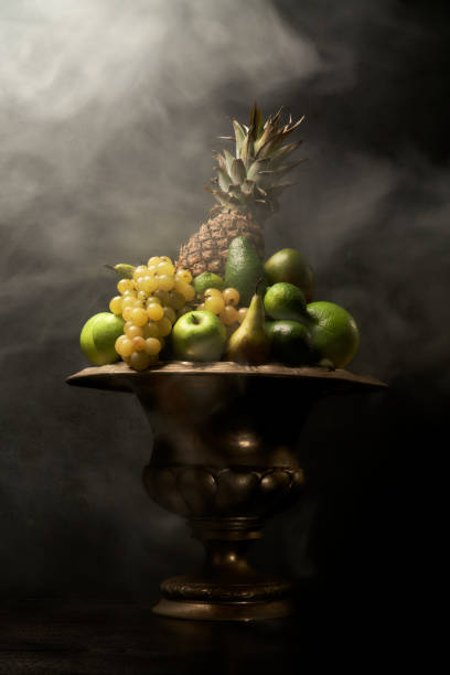 Still Life with Green Fruits in a dark smoky room Still Life with many different tropical green fruits in big silver vase / bowl. Dark wall background, antique wooden table, pineapple, wine grapes, green apples, limes. Great for designers and interior decoration. antique fruit bowl stock pictures, royalty-free photos & images