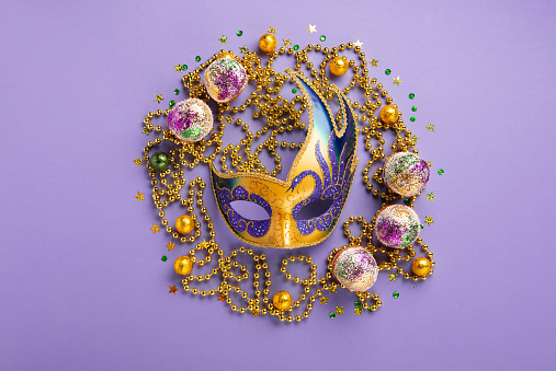 Mardi Gras King Cake cupcake or muffins, masquerade festival carnival mask, gold beads and golden, green confetti on purple background. Holiday party invitation, greeting card concept.