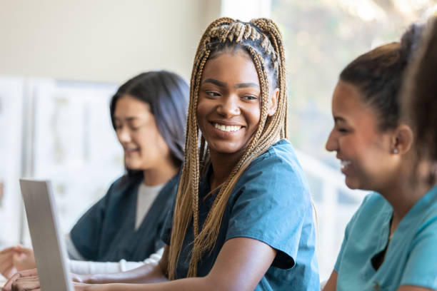 young adult nursing or medical student wearing scrubs uses laptop computer and discusses medical training class assignment with classmate - mature student learning training computer imagens e fotografias de stock