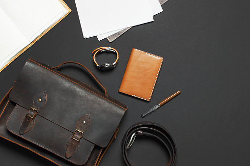Fashionable concept. Brown leather men's bag, wristwatch, leather passport cover, pen, blank white sheets on black background top view flat lay with copy space Accessories businessman stylish clothes