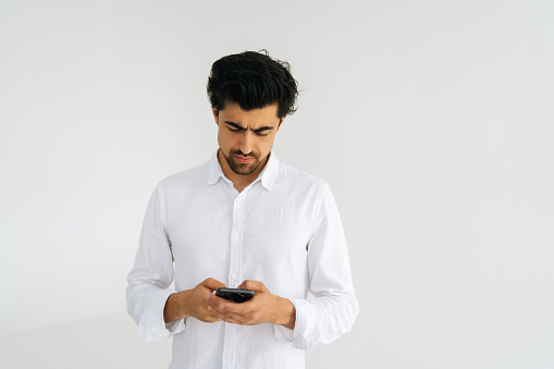 Studio portrait of serious bearded young man in shirt holding smartphone, using online mobile apps, looking on device screen, chatting in messengers standing on white isolated background.