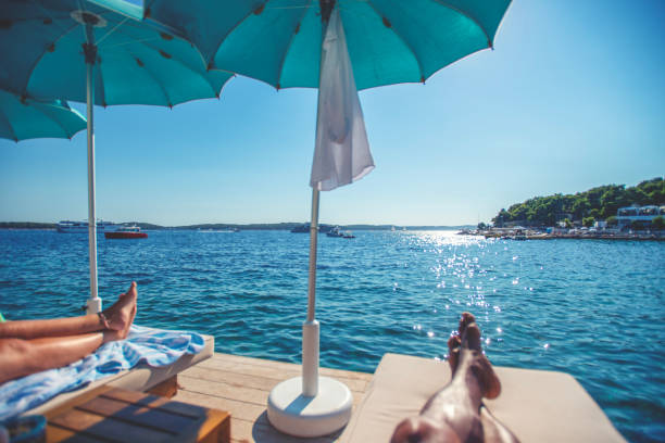 Couple lying by the ocean on deck chairs. Only their feet can be seen with the ocean in the background.