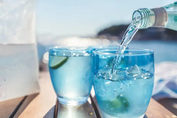 Pouring water from a bottle into a glass at the beach. Lemon and ice in the blue glasses. Backlit at sunset