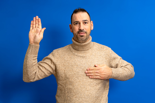 Bearded hispanic man in turtleneck with hand on chest making an oath of honor, swears to tell the whole truth without fear of consequences. Isolated on blue background