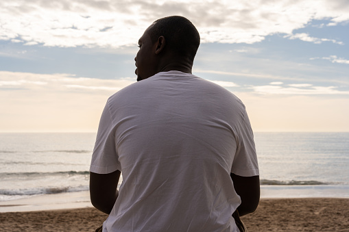 Back view of African American male in white casual clothes looking away while sitting on sandy beach against cloudy sky during sundown