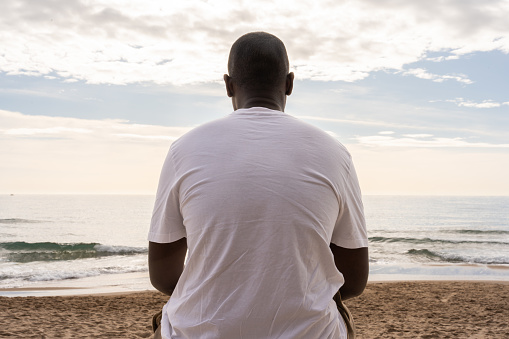 Low angle back view of unrecognizable African American male in casual white t shirt sitting on sandy beach and admiring waving sea