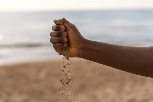 Crop anonymous African American male pouring sand from hand during day on beach near waving sea