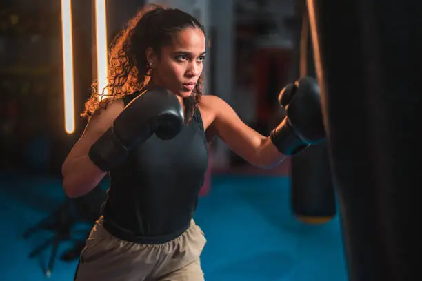 Serious young Latin American female boxer punching a boxing bag. She is exercising and training with boxing equipment at a fitness center.