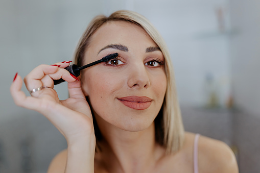 A close-up of a young Caucasian woman looking at the camera and applying mascara with a smile on her face.