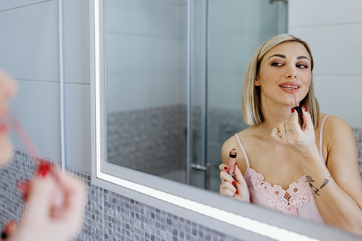 A young Caucasian woman is looking at her reflection in the mirror and applying lip gloss.