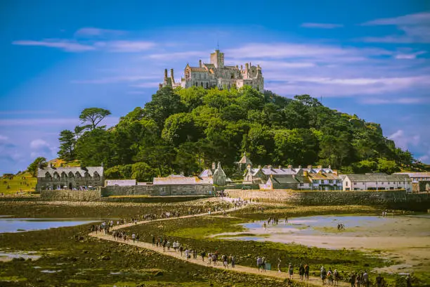 View of long line of people walking to St. Michael's Mount island during low tide in summer; blue sky with light clouds in background