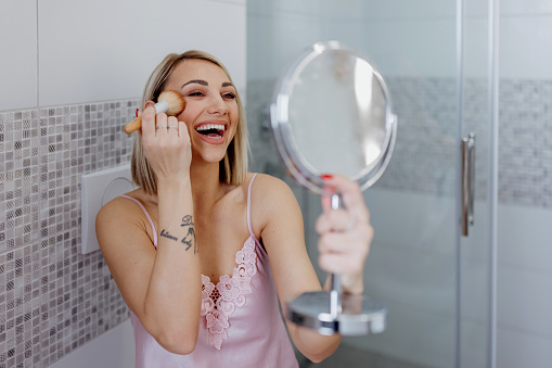 A young Caucasian woman is having fun and laughing while applying blush in her bathroom.