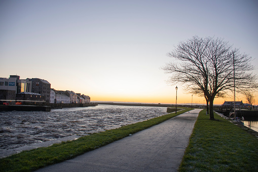 sunrise on the river corrib galway with frozen sidewalk and dry golden hour tree