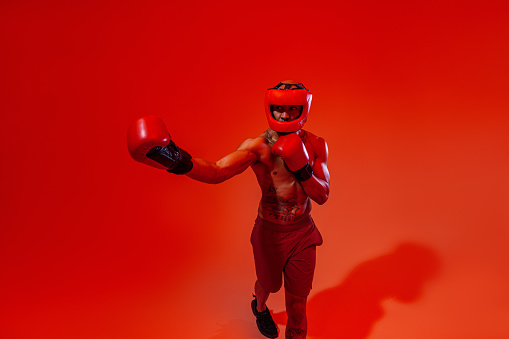 Man wearing head guard and boxing gloves practicing for fight on studio background with color filter