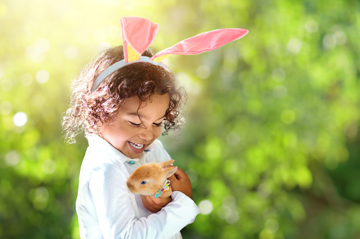 Little curly boy with Easter bunny. Child playing with rabbit pet. Kid in bunny ears on Easter egg hunt. Spring holiday celebration in sunny garden. Adorable baby with pet.
